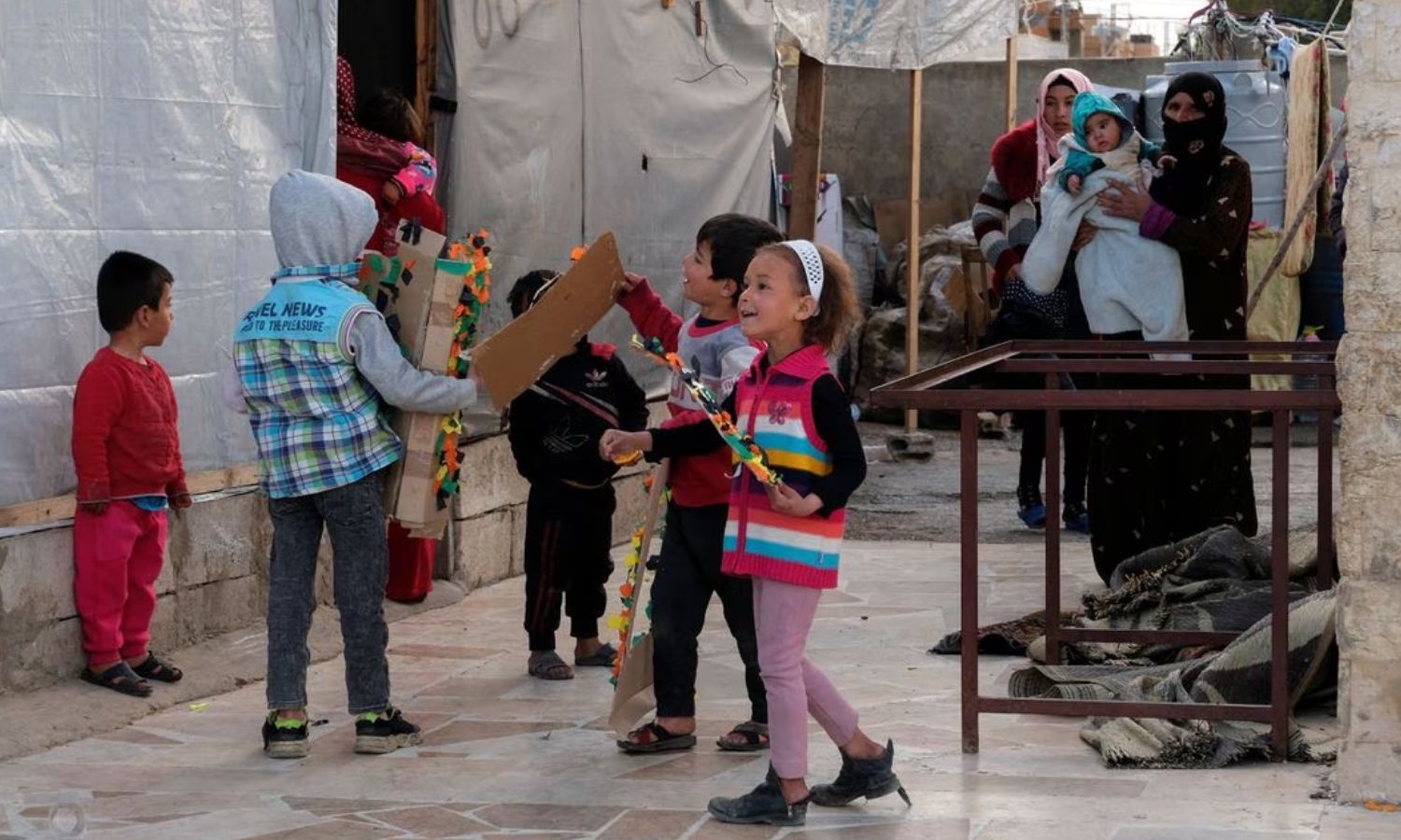 سيّدة تحمل طفلًا تقف قرب مجموعة أطفال يلعبون أمام خيمة للاجئين سوريين في منطقة البقعة التابعة للبقاع في لبنان- 5 من نيسان 2023 (رويترز)
