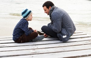 Father and son (10-11) sitting at end of dock at edge of lake, talking, side view