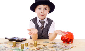 Sly boy in black hat with empty hands at the table with pile of money, isolated on white