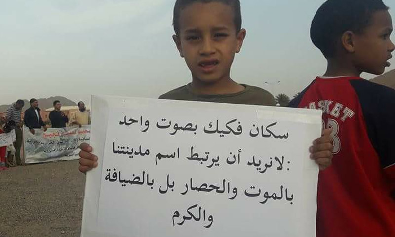 طفل مغربي يحمل لافتة احتجاجًأ على إهمال سوريين عالقين على الحدود مع الجزائر منذ نيسان 2017 (فيس بوك)