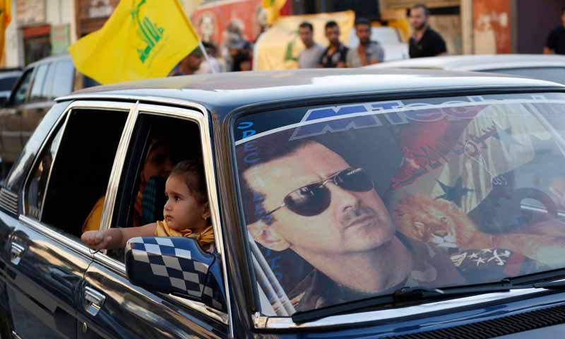 صورة لرئيس النظام السوري بشار الأسد على زجاج سيارة وورائه راية لحزب الله اللبناني أثناء احتفالية بعيد الجيش السوري - (رويترز)