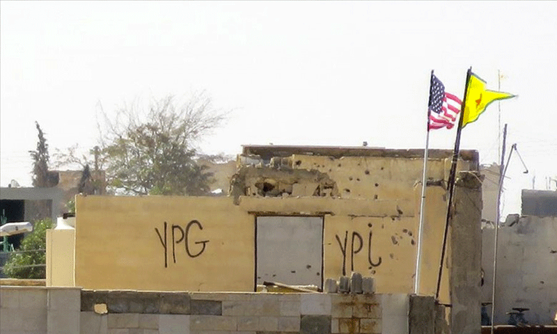 علم وحدات حماية الشعب الكردية إلى جانب العلم الأمريكي في بلدة تل أبيض شمال سوريا (الأناضول)