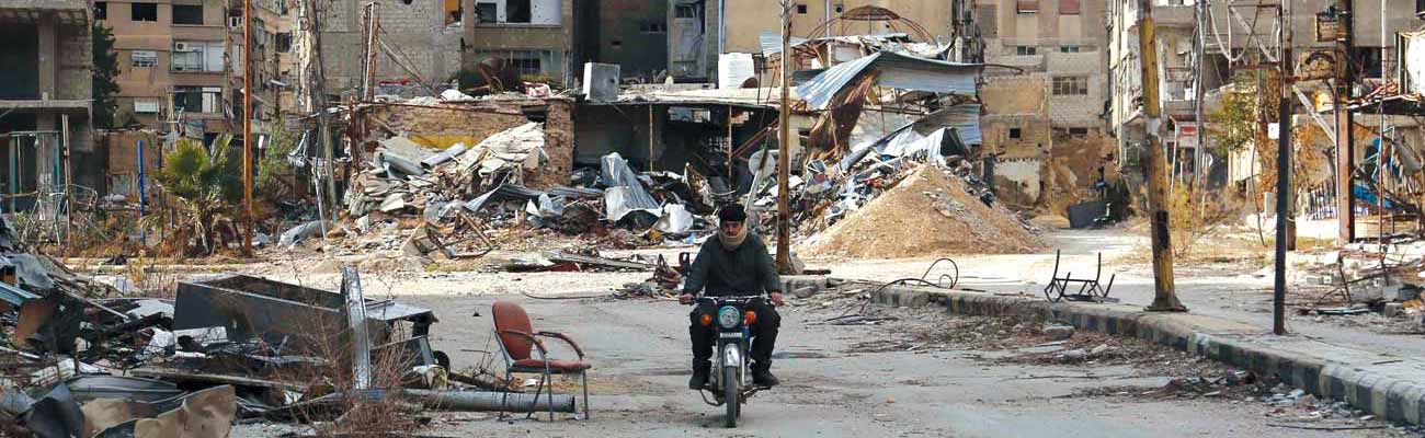 رجل يقود دراجته النارية خلف المباني المدمرة في مدينة حرستا شرقي دمشق. (AFP)