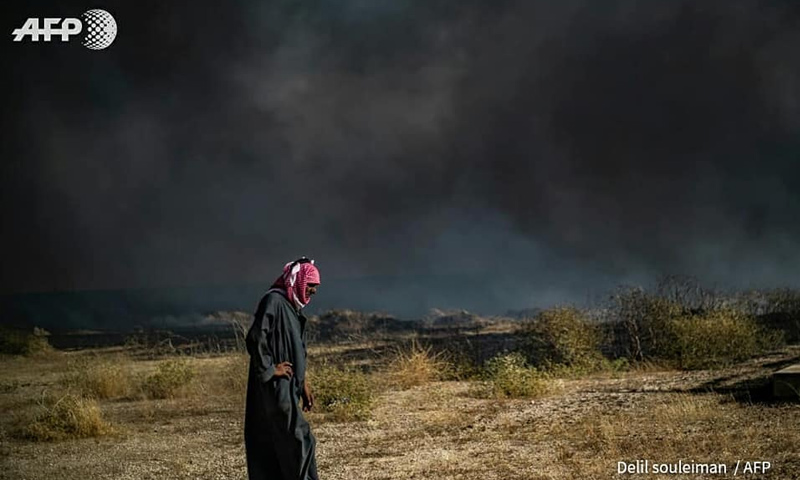 رجل يسير في حقل بينما يتصاعد الدخان في حقل زراعي في بلدة القحطانية في محافظة الحسكة- 10 من حزيران 2019 (AFP)