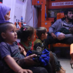 رجل وامرأة يحضنان ابنيهما وبجوارهم أطفال مصابين بتسمم في سيارة إسعاف نتيجة تناول طعام فاسد مقدم من أحد الجمعيات الخيرية في مخيم رعاية الطفولة شمال إدلب - 11 أيار 2020 (عنب بلدي)