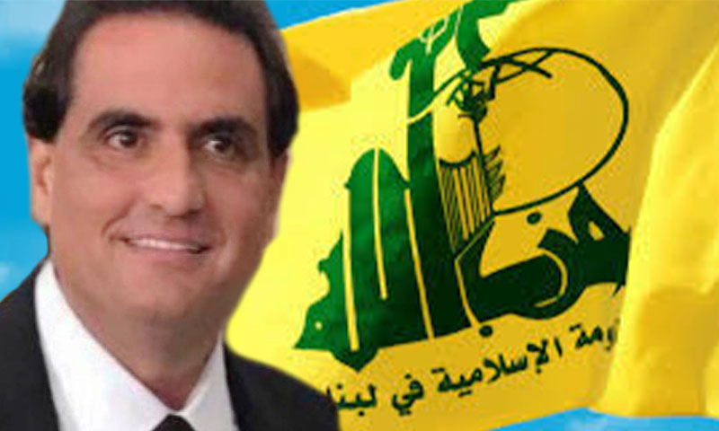 أليكس صعب وخلفه علم حزب الله اللبناني (تعديل عنب بلدي)
