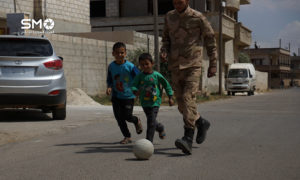 طفلان يلعبان بالكرة مع أحد عناصر الجيش الجر في درعا – أيار 2016 (الهيئة السورية للإعلام)