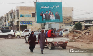 أشخاص يمشون في الشارع في مدينة الرقة وتظهر لافتة تحث الناس على الاهتمام بالوقاية من فيروس 