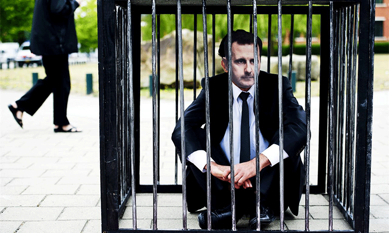متظاهر يرتدي قناع وجه رئيس النظام السوري بشار الأسد جالسًا على الأرض داخل قفص الاتهام أمام “المحكمة الجنائية الدولية” في لاهاي غربي هولندا- 7 من حزيران 2011 (ROBIN UTRECHT / ANP / AFP)
