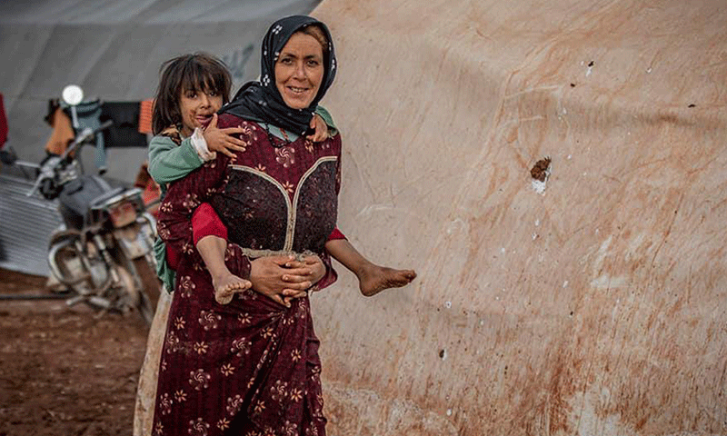 لاجئة سورية تحمل طفلتها في مخيم بمدينة سرمدا شمالي محافظة إدلب- 2019 (الشبكة السورية لحقوق الإنسان)