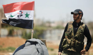   مجند في جيش النظام السوري بجوار علم يحمل صورة رئيس النظام بشار الأسد. درعا - تموز 2018 (رويترز)
