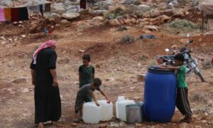 رجل يقف مع ثلاثة أطفال إلى جانب برميل أزرق وعبوات بلاستيكية بيضاء مملوءة بالماء في أحد المخيمات_ 26 من أيار 2022 (منسقو استجابة سوريا/ فيس بوك)