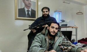 ضابط المخابرات العسكرية أمجد يوسف يجلس أمام طاولة وخلفه زميل له يحمل البندقية في مكتب داخل فرع المنطقة 