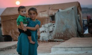 طفلة سورية في أحد مخيمات الشمال السوري تحمل بيديها طفلًا صغيرًا وقت غروب الشمس_ 4 من حزيران 2022 (الدفاع المدني السوري/ فيس بوك)