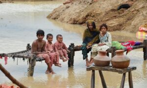 امرأة وأطفال يجلسون على أسِرَّة من الخشب والحبال موضوعة فوق مياه الفيضانات إلى جانب منزلهم المدمر بعد هطول الأمطار في ديرا الله يار التابعة لإقليم بلوشستان_ 25 من آب 2022 (رويترز)