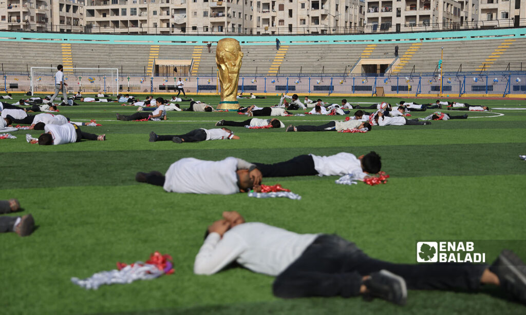رجال وشباب مستلقون على الأرض خوفًا من القصف وخلفهم شعار كأس العالم في مشهد تمثيلي ضمن افتتاحية ملعب إدلب البلدي - 20 تشرين الثاني 2022 (عنب بلدي \ أنس الخولي )