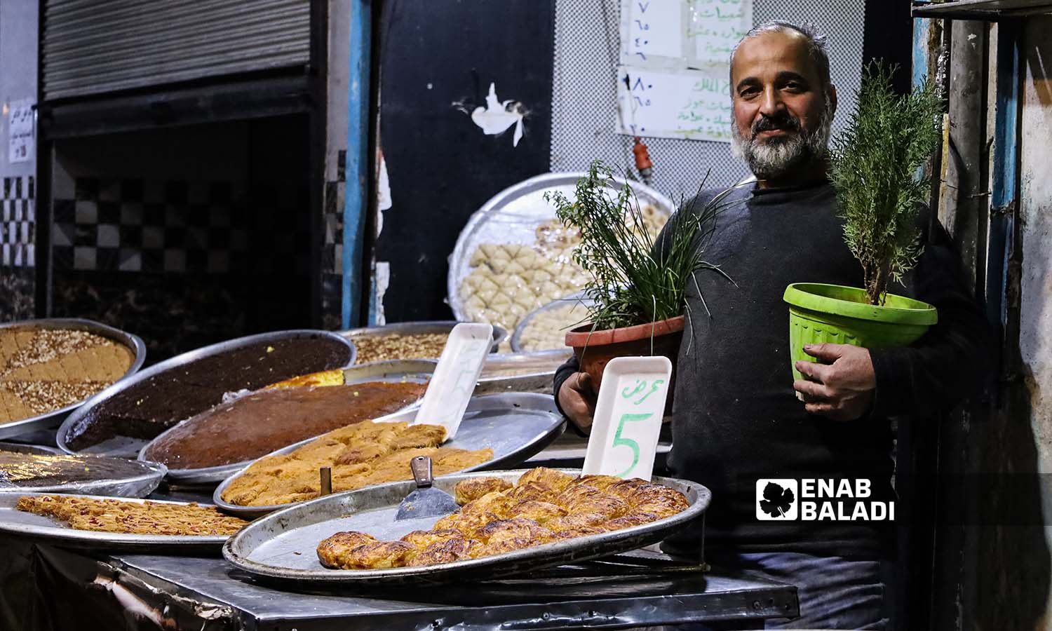 بائع حلويات يمسك بيديه أحواض نباتات بالقرب من طاولة عرض بضائعه في مدينة إدلب- 25 من تشرين الثاني 2022 (عنب بلدي/ مجد هامو)