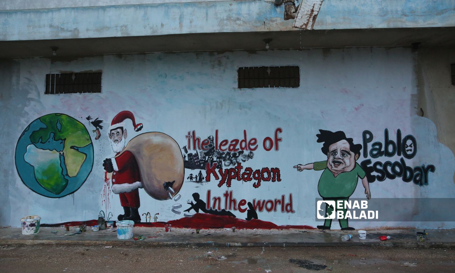 الرسام عزيز أسمر وأنيس حمدون، يرسمان جدارية بمناسبة رأس السنة الميلادية، تظهر بشار الأسد بشخصية بابا نويل وهو يوزع حبوب الكبتاجون في مدينة بنش - 25 كانون الأول 2022 (عنب بلدي / أنس الخولي)