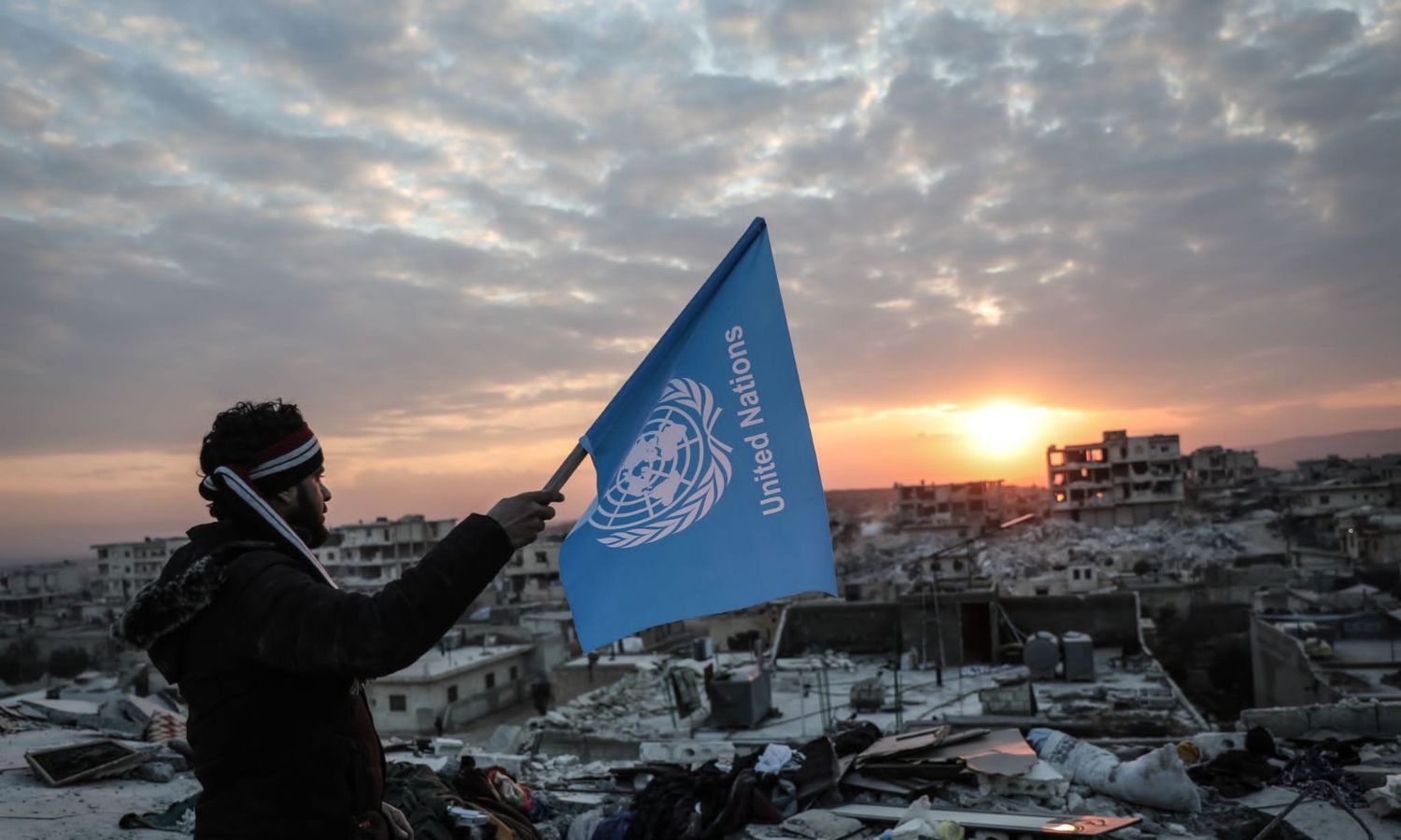 شاب يحمل علم الأمم المتحدة وهو مقلوب أعلى ركام بناء مدمّر في بلدة جنديرس بريف حلب الشمالي استنكارًا لتأخر الاستجابة إثر زلزال ضرب مناطق تركيا وأربع سورية- 11 شباط 2023 (متداول/ وليد أكتع)