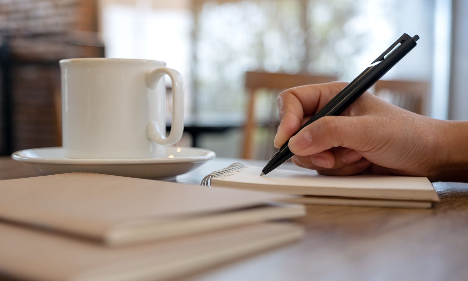 تعبيرية- شخص يمسك بقلم ويكتب على دفتر يجاوره فنجان قهوة
