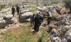 متطوعان في "الدفاع المدني السوري" وأهالي ينقلون جثة شخص قُتل بطيران مسيّر أمريكي على أطراف بلدة قورقانيا شمالي إدلب- 3 من أيار 2023 (الدفاع المدني / فيس بوك)
