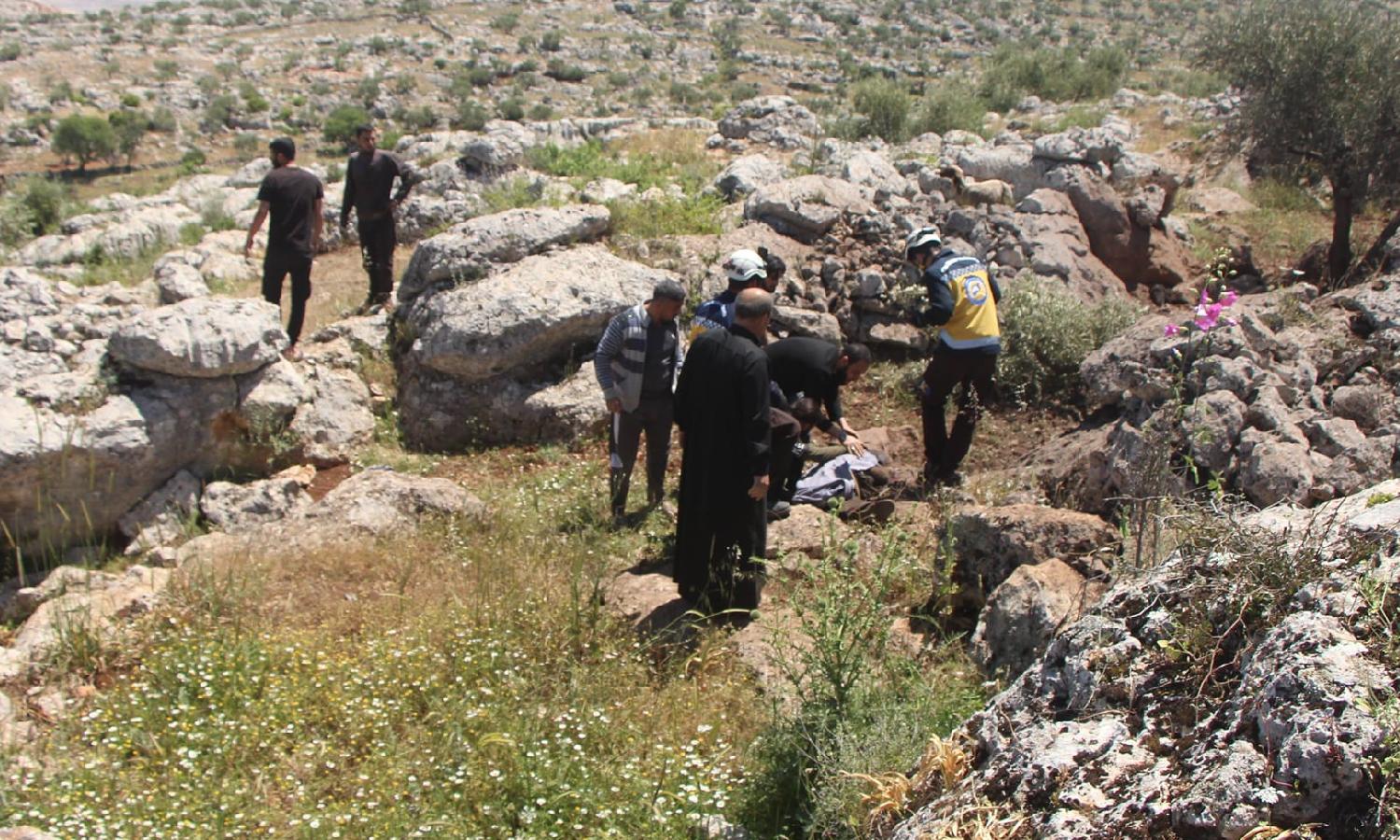 متطوعان في "الدفاع المدني السوري" وأهالي ينقلون جثة شخص قُتل بطيران مسيّر مجهول الهوية على أطراف بلدة قورقانيا شمالي إدلب- 3 من أيار 2023 (الدفاع المدني / فيس بوك)