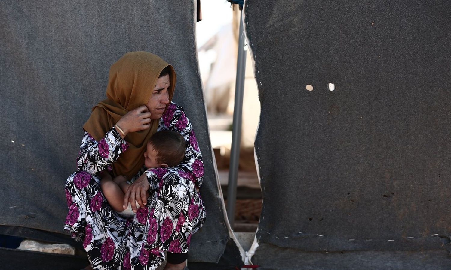 امرأة تحمل طفلها وتجلس قرب خيمتها في أحد مخيمات إدلب (رويترز)