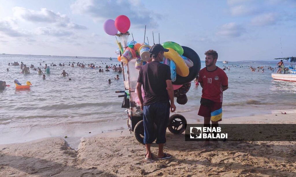 يسعى البائعون الجوالون لجلب رزقهم من خلال تجولهم في شاطئ "الكرنك" لبيع المرطبات ومستلزمات البحر - 12 من آب 2023 (عنب بلدي/ليندا علي)