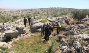 متطوعان في "الدفاع المدني السوري" وأهالي ينقلون جثة شخص قُتل بطيران مسيّر أمريكي على أطراف بلدة قورقانيا شمالي إدلب- 3 من أيار 2023 (الدفاع المدني / فيس بوك)