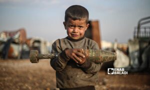 يحمل الطفل قذيفة مدفعية غير منفجرة في ساحة لتجميع الصواريخ في ريف إدلب الشمالي بين ذراعيه وهو يعمل مع عائلته على تأمين رزقهم- 5 من آذار 2021 (عنب بلدي/ يوسف غريبي)