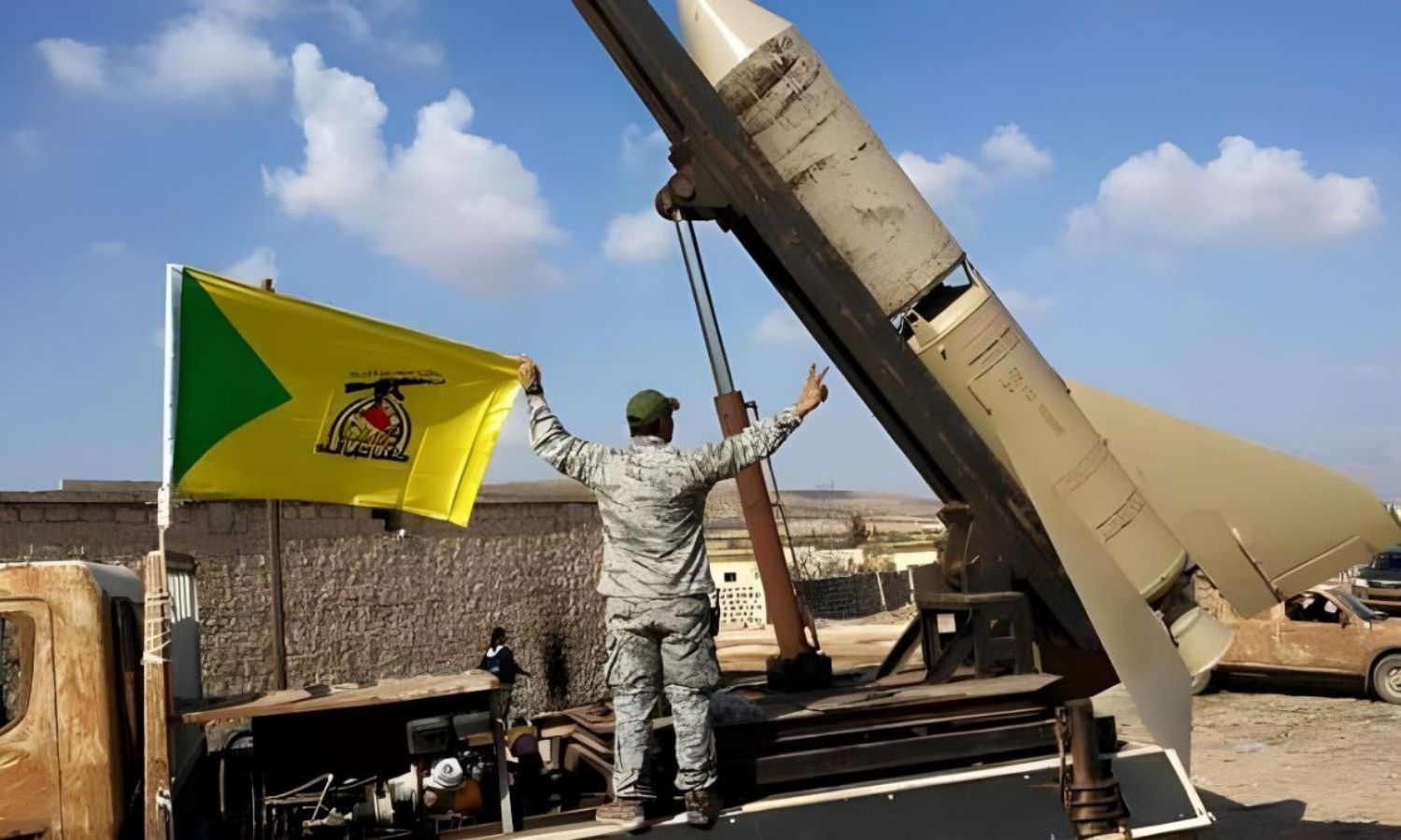 مقاتل بجانب منصة إطلاق صواريخ يرفع علم كتائب حزب الله العراقية (الموقع الرسمي لكتائب حزب الله)