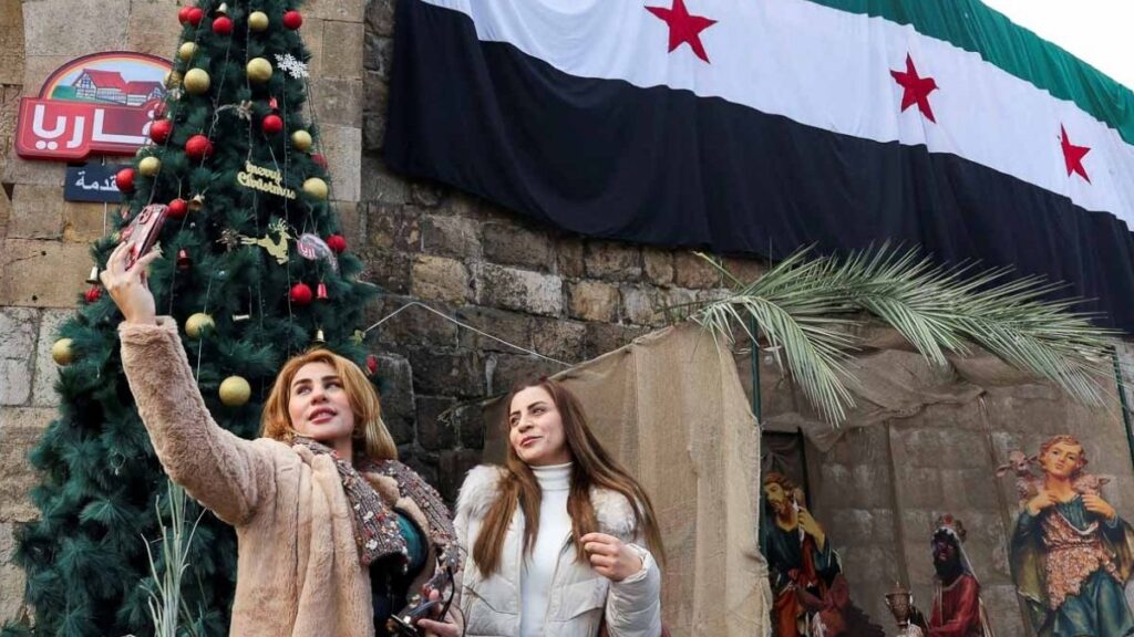 نساء يستخدمن هواتفهن المحمولة أمام علم في ساحة باب توما ليلة رأس السنة 31 كانون الأول 2024 (رويترز)