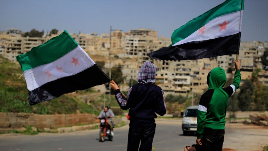أطفال يرفعون العلم السوري خلال التجهيز لانطلاق مظاهرة تحاكي المظاهرة الأولى التي انطلقت في درعا - 18 آذار 2026 (عنب بلدي)