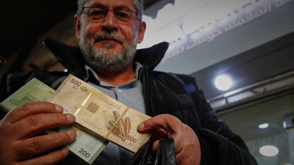 رجل يعرض أوراقًا نقدية من فئة 500 و200 ليرة سورية جديدة أمام مكتب صرافة في دمشق - 3 كانون الثاني 2026 (AP/عمر صناديقي)