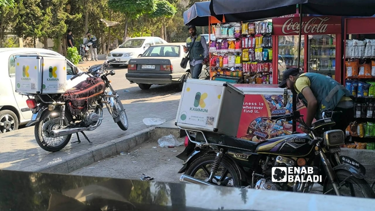 عاملان في توصيل الطلبات يقفان إلى جانب دراجتيهما أمام متجر في دمشق بانتظار طلبات جديدة - 16 تموز 2026 (عنب بلدي)