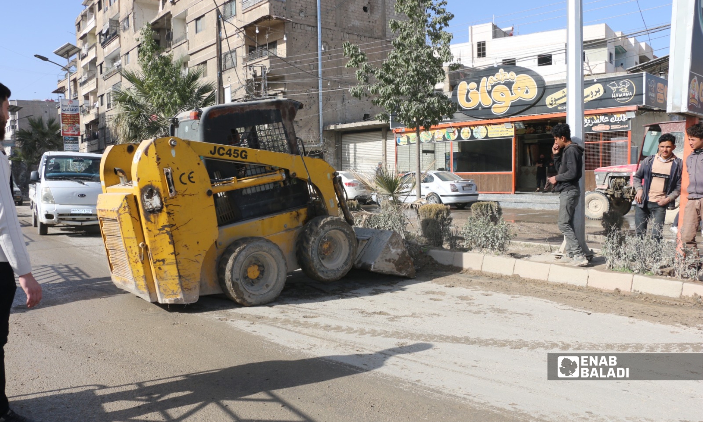 عمال يزيلون أنقاضًا وردمًا من رصيف أحد الشوارع في مدينة داريا باستخدام أدوات يدوية وعربة نقل، ضمن حملة تنظيف خدمية بالتعاون بين UNDP وبلدية داريا - 5 شباط 2026 (عنب بلدي)