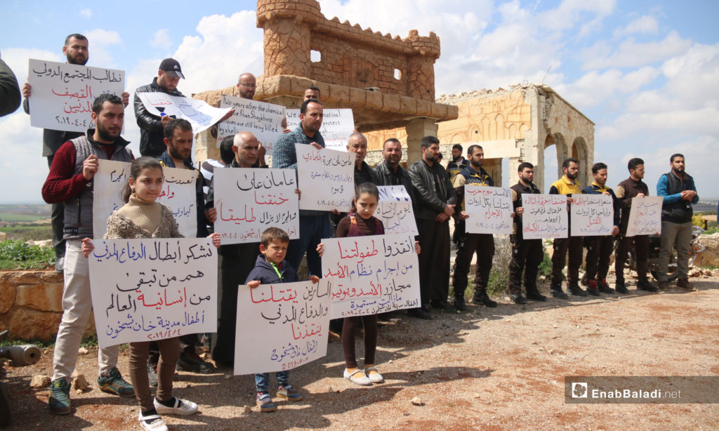A vigil at the second anniversary of the chemical massacre of Khan Shaykhun, rural Idlib – April 4, 2019 (Enab Baladi) The signs held by the people say: “Suffocated, burnt and mutilated, are there any death methods left that you have not tried yet; hold al-Assad accountable, chemical weapons Khan Shaykhun/ Al-Assad is a war criminal, killing us with all the tools of crime/ two years ago, he suffocated us, and he is yet killing us/ 04.04.2017 is a day that our generations will not forget/ Bashar al-Assad is killing his people with an international license/ Two years have passed since the chemical weapons’ massacre and the massacres of the Russians and the regime are yet going on/We demand that the international community stop the shelling and protect the civilians/ Save our childhood from the criminality of the al-Assad regime; Massacres by al-Assad and Putin are continuing/ Two years have passed since we were suffocated and the criminal is still free/The tools are many and the criminal is but one; chemical weapons, aircraft, missiles, and the massacres are still going on/We thank the heroes of the Civil Defense, they are what is left of the world’s humanity, the children of the city of Khan Shaykhun/He is making an art out of killing his people and has a plan to get back Golan/Two years have passed since our suffocation, and our children are still suffering the bitterness of daily shelling.”