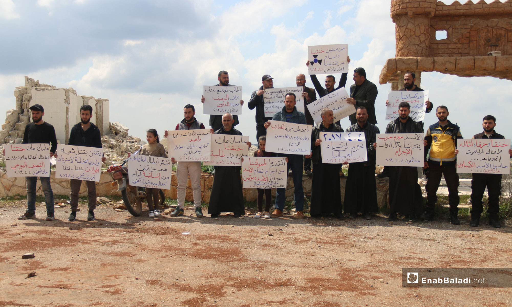A vigil at the second anniversary of the chemical massacre of Khan Shaykhun, rural Idlib – April 4, 2019 (Enab Baladi)
The signs held by the people say: “Suffocated, burnt and mutilated, are there any death methods left that you have not tried yet; hold al-Assad accountable, chemical weapons  Khan Shaykhun/ Al-Assad is a war criminal, killing us with all the tools of crime/ two years ago, he suffocated us, and he is yet killing us/ 04.04.2017 is a day that our generations will not forget/ Bashar al-Assad is killing his people with an international license/ Two years have passed since the chemical weapons’ massacre and the massacres of the Russians and the regime are yet going on/We demand that the international community stop the shelling and protect the civilians/ Save our childhood from the criminality of the al-Assad regime; Massacres by al-Assad and Putin are continuing/ Two years have passed since we were suffocated and the criminal is still free/The tools are many and the criminal is but one; chemical weapons, aircraft, missiles, and the massacres are still going on/We thank the heroes of the Civil Defense, they are what is left of the world’s humanity, the children of the city of Khan Shaykhun/He is making an art out of killing his people and has a plan to get back Golan/Two years have passed since our suffocation, and our children are still suffering the bitterness of daily shelling.”
