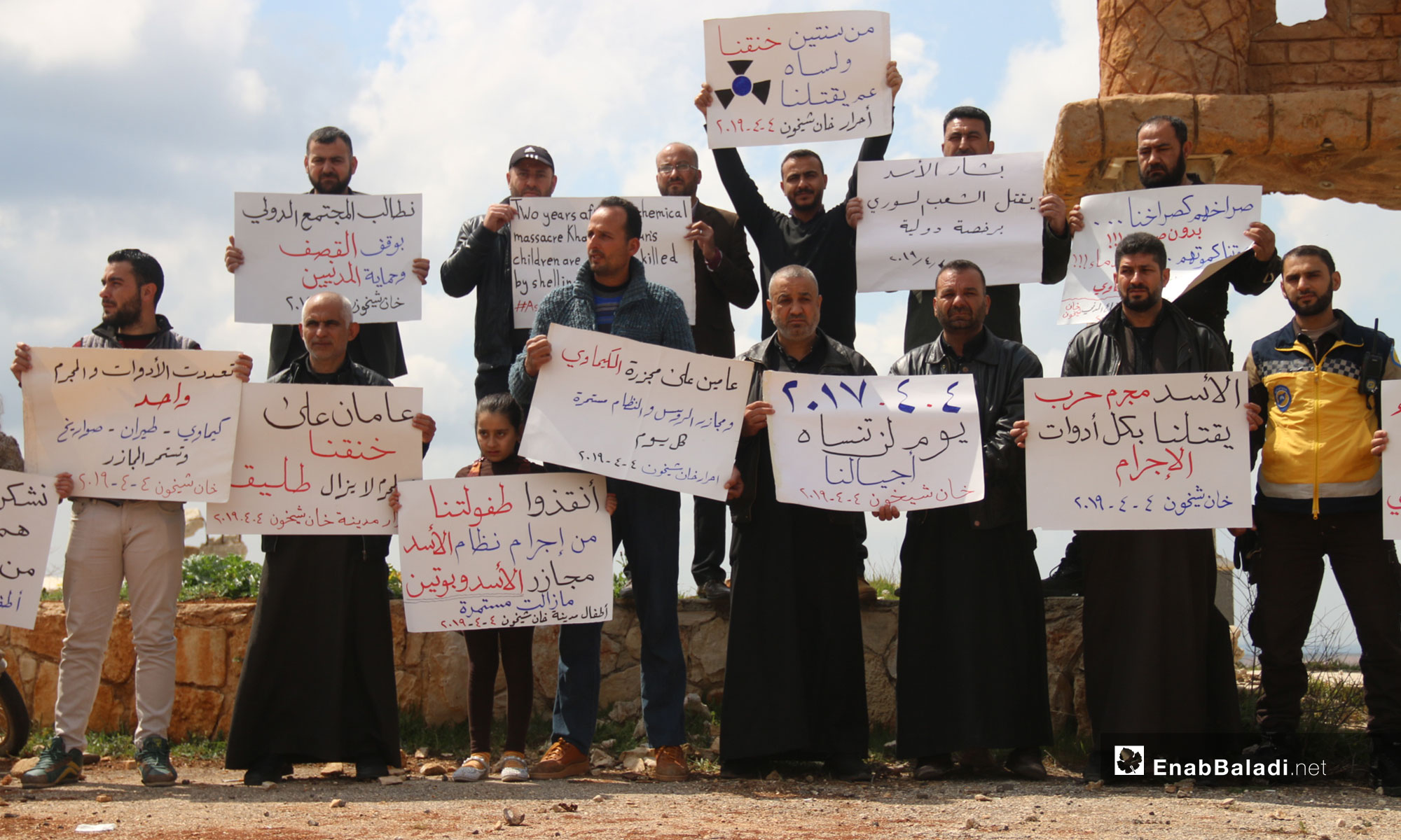 A vigil at the second anniversary of the chemical massacre of Khan Shaykhun, rural Idlib – April 4, 2019 (Enab Baladi)
The signs held by the people say: “Suffocated, burnt and mutilated, are there any death methods left that you have not tried yet; hold al-Assad accountable, chemical weapons  Khan Shaykhun/ Al-Assad is a war criminal, killing us with all the tools of crime/ two years ago, he suffocated us, and he is yet killing us/ 04.04.2017 is a day that our generations will not forget/ Bashar al-Assad is killing his people with an international license/ Two years have passed since the chemical weapons’ massacre and the massacres of the Russians and the regime are yet going on/We demand that the international community stop the shelling and protect the civilians/ Save our childhood from the criminality of the al-Assad regime; Massacres by al-Assad and Putin are continuing/ Two years have passed since we were suffocated and the criminal is still free/The tools are many and the criminal is but one; chemical weapons, aircraft, missiles, and the massacres are still going on/We thank the heroes of the Civil Defense, they are what is left of the world’s humanity, the children of the city of Khan Shaykhun/He is making an art out of killing his people and has a plan to get back Golan/Two years have passed since our suffocation, and our children are still suffering the bitterness of daily shelling.”
