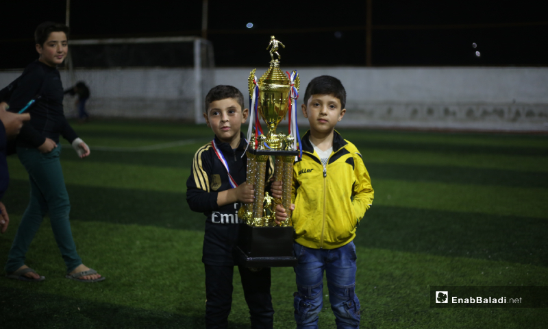 Two children carrying the North Stars League Championship Cup after the end of the final match, with the winning of Akrabat team over the Deir Hassan team in Idlib – 03 May 2020 (Enab Baladi)