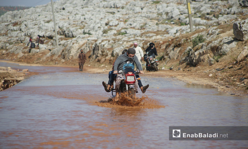 Two men attempting to cross the stormwater pond by a motorcycle after the rainstorm that hit “Sahl al-Khair” camp for Syrian internally displaced people (IDPs) near Kafr Bunni in northern Idlib countryside – 19 June 2020 (Enab Baladi / Yousef Ghuraibi)