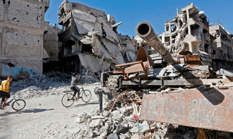 Two boys riding their bicycles in front of a tank and destroyed buildings in the Yarmouk camp in 2018 (The Daily Star)