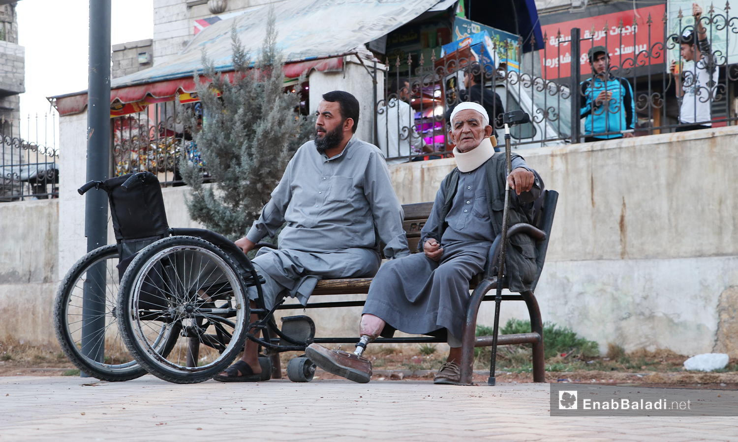 An old man with his crutch and prosthetic limb is resting on a public wooden chair next to a man whose leg is amputated and uses a wheelchair - 24 November 2020 (Enab Baladi / Asim Melhem)