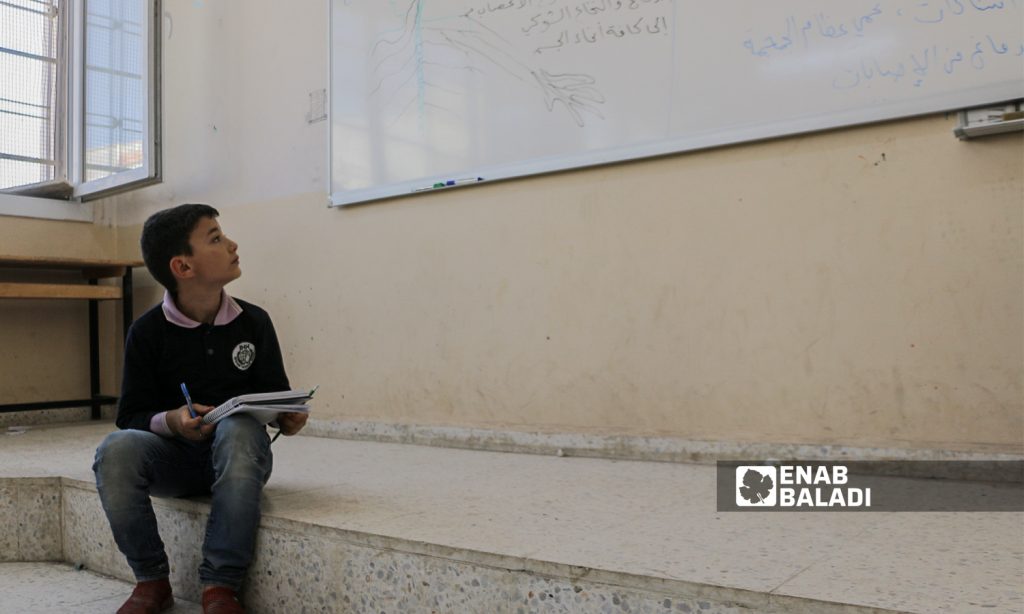 A primary schooler is sitting on the ground next to the whiteboard writing down his notes in a classroom in Azaz- 22 September 2021 (Enab Baladi- Walid Othman)