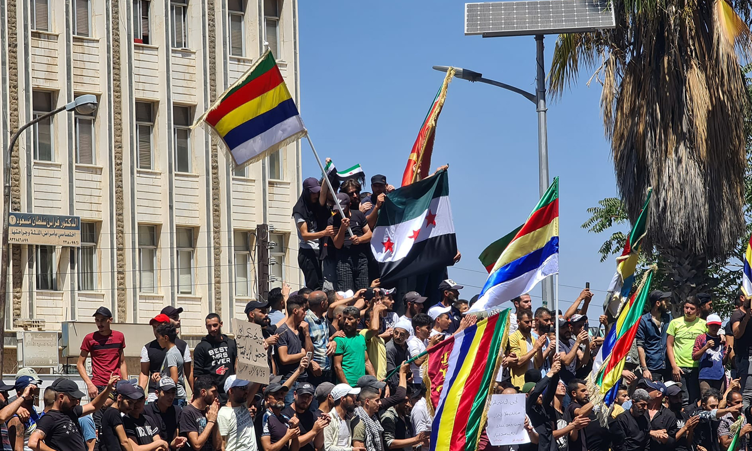 Residents of the southern city of As-Suwayda during protests in the newly-named al-Karamah (Dignity) Square raise the flag of the Syrian revolution next to the five-colored flag of the Druze community - August 24, 2023 (Suwayda 24)