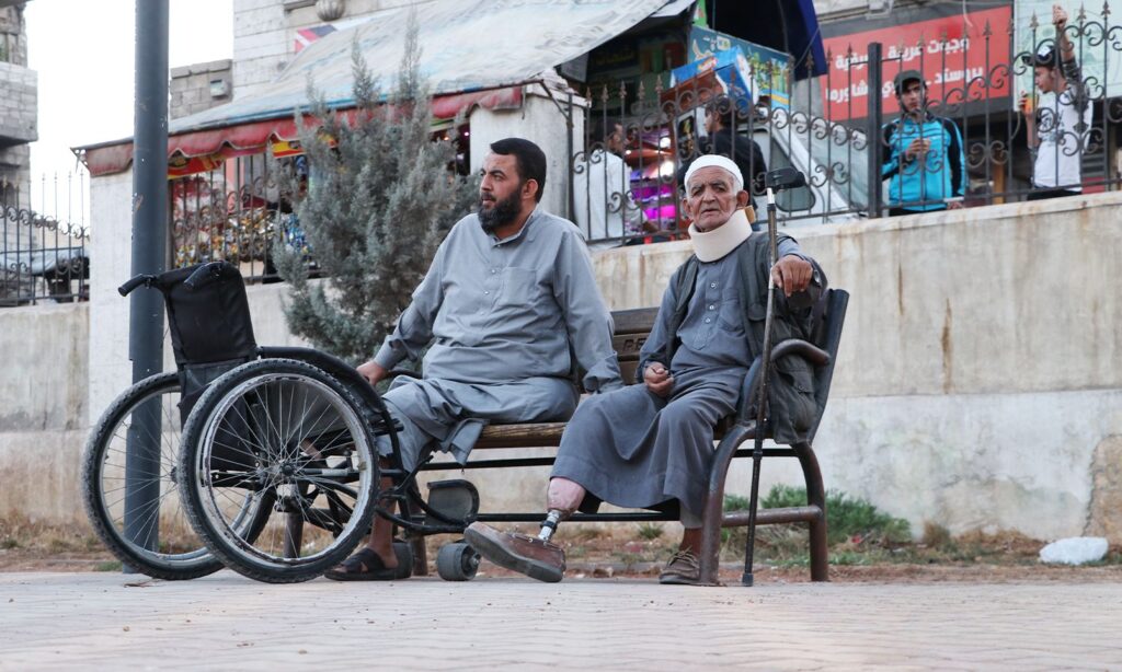 An elderly man leaning on his prosthetic leg, sitting on a wooden bench near an amputee facing a wheelchair - November 24, 2020 (Enab Baladi/Asim Melhem)