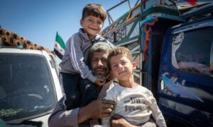A man carries two children before leaving a displacement camp in northern Syria, September 27. (OCHA)